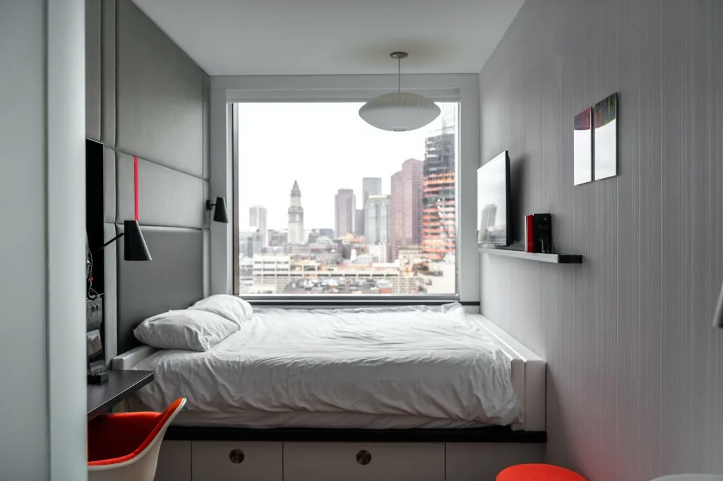 Very small bedroom with muted colors and red accents. Window at the end of the room shows downtown view.