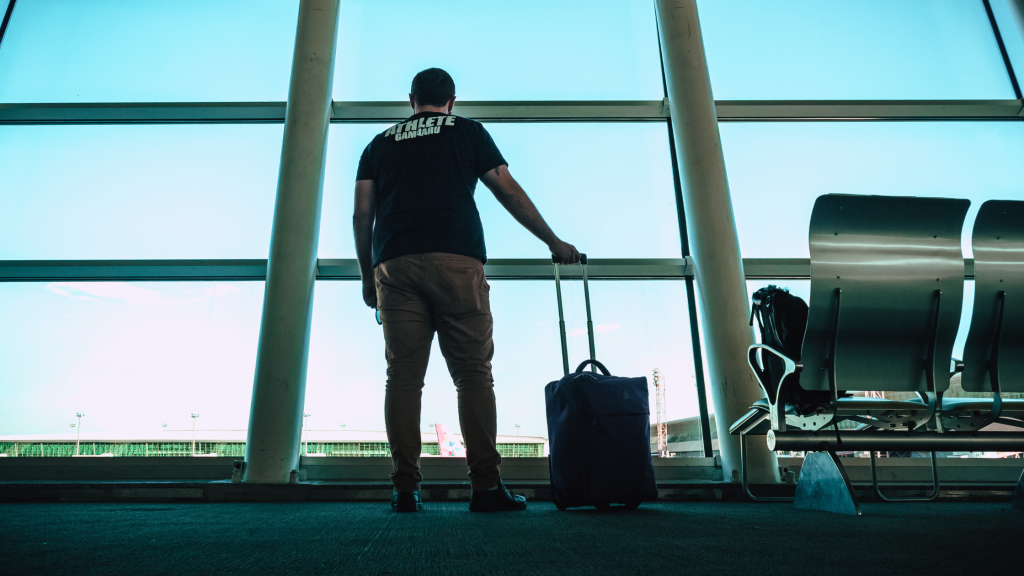 A man watching an airport window while holding to his luggage.