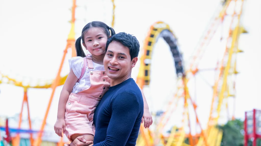 An image of a father and daughter posing for a photo in front of a roller coaster.