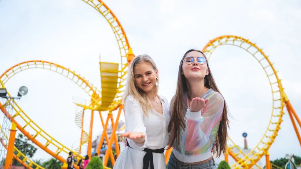 Two female visitors of an amusement park in front of a roller coaster.