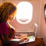 A woman using her laptop onboard an airplane, seated near the window with natural light streaming in. Ideal for an article on travel rules and tips, answering the question, Can I bring my laptop in my carry-on?