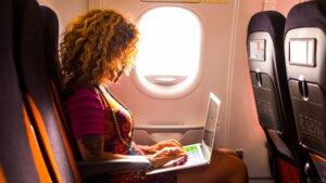 A woman using her laptop onboard an airplane, seated near the window with natural light streaming in. Ideal for an article on travel rules and tips, answering the question, Can I bring my laptop in my carry-on?