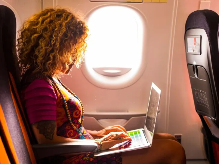 A woman using her laptop onboard an airplane, seated near the window with natural light streaming in. Ideal for an article on travel rules and tips, answering the question, Can I bring my laptop in my carry-on?