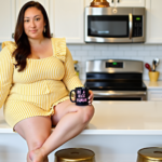 A woman in a yellow striped dress sits on a kitchen counter in a modern kitchen, holding a black mug with the phrase "Be A Nice Human" in pink letters. The setting suggests a cozy, well-maintained home, emphasizing a warm and welcoming ambiance that could appeal to potential Airbnb guests in a primary residence setup
