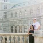 A couple stands together on a stone balcony in front of a grand historic building, the man looking off into the distance while the woman leans into him, conveying unity and partnership. This image captures the essence of building a supportive relationship in marriage, aligning with the theme of fostering mutual understanding and getting your husband on your side.