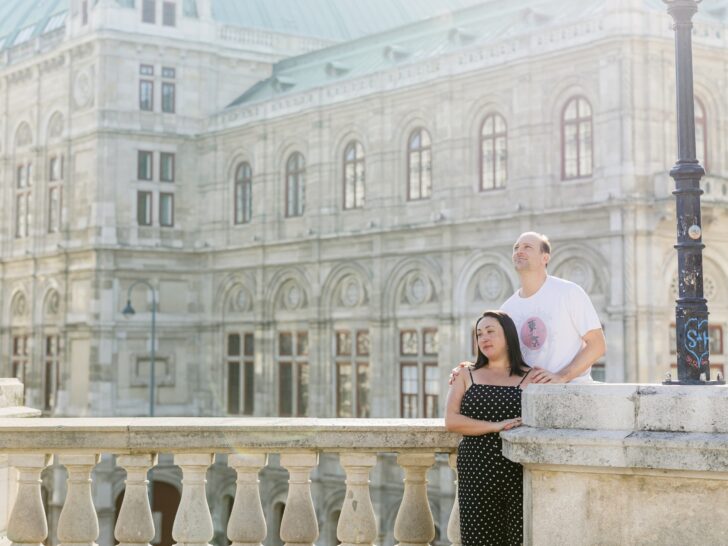 A couple stands together on a stone balcony in front of a grand historic building, the man looking off into the distance while the woman leans into him, conveying unity and partnership. This image captures the essence of building a supportive relationship in marriage, aligning with the theme of fostering mutual understanding and getting your husband on your side.