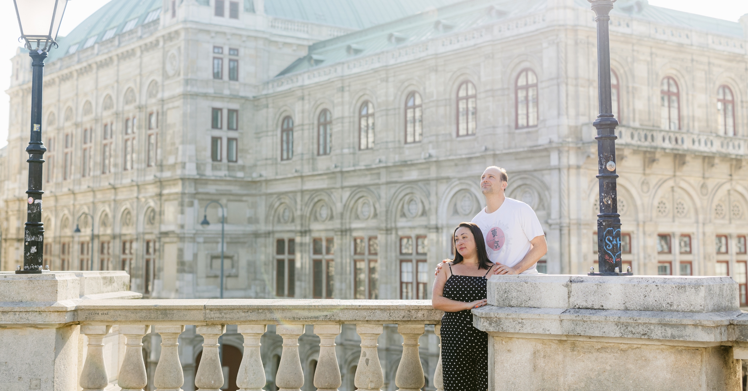 A couple stands together on a stone balcony in front of a grand historic building, the man looking off into the distance while the woman leans into him, conveying unity and partnership. This image captures the essence of building a supportive relationship in marriage, aligning with the theme of fostering mutual understanding and getting your husband on your side.