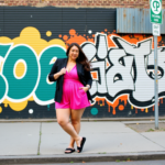 A woman in a vibrant pink romper and black blazer stands confidently in front of a colorful graffiti wall, exuding self-assurance. Her relaxed stance and direct gaze reflect an empowered attitude, aligning with themes of overcoming self-doubt and embracing self-confidence.