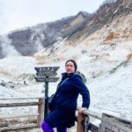 A woman standing on a wooden walkway with snow-covered geothermal terrain and steaming hills in the background, next to a sign that reads 'Danger: Keep Out.' Perfect for an article on motivational quotes about struggle and overcoming challenges.