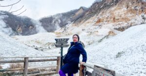 A woman standing on a wooden walkway with snow-covered geothermal terrain and steaming hills in the background, next to a sign that reads 'Danger: Keep Out.' Perfect for an article on motivational quotes about struggle and overcoming challenges.