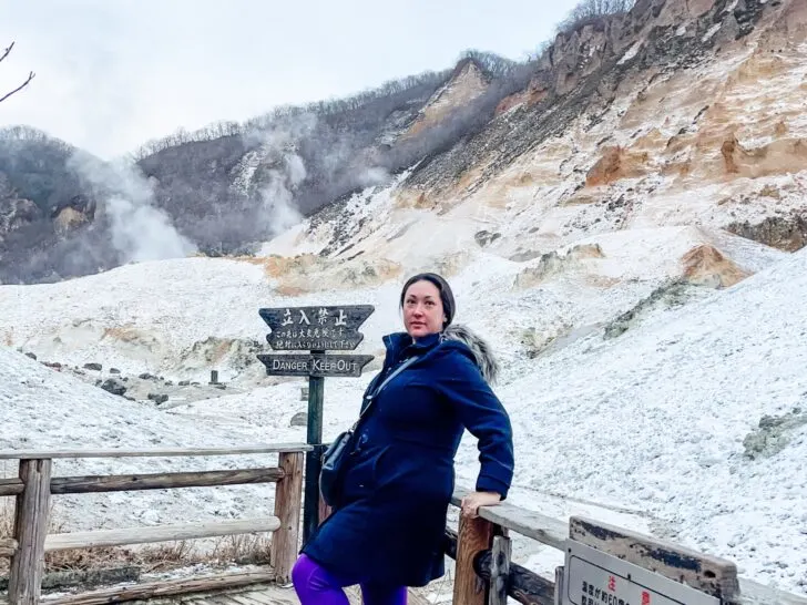 A woman standing on a wooden walkway with snow-covered geothermal terrain and steaming hills in the background, next to a sign that reads 'Danger: Keep Out.' Perfect for an article on motivational quotes about struggle and overcoming challenges.