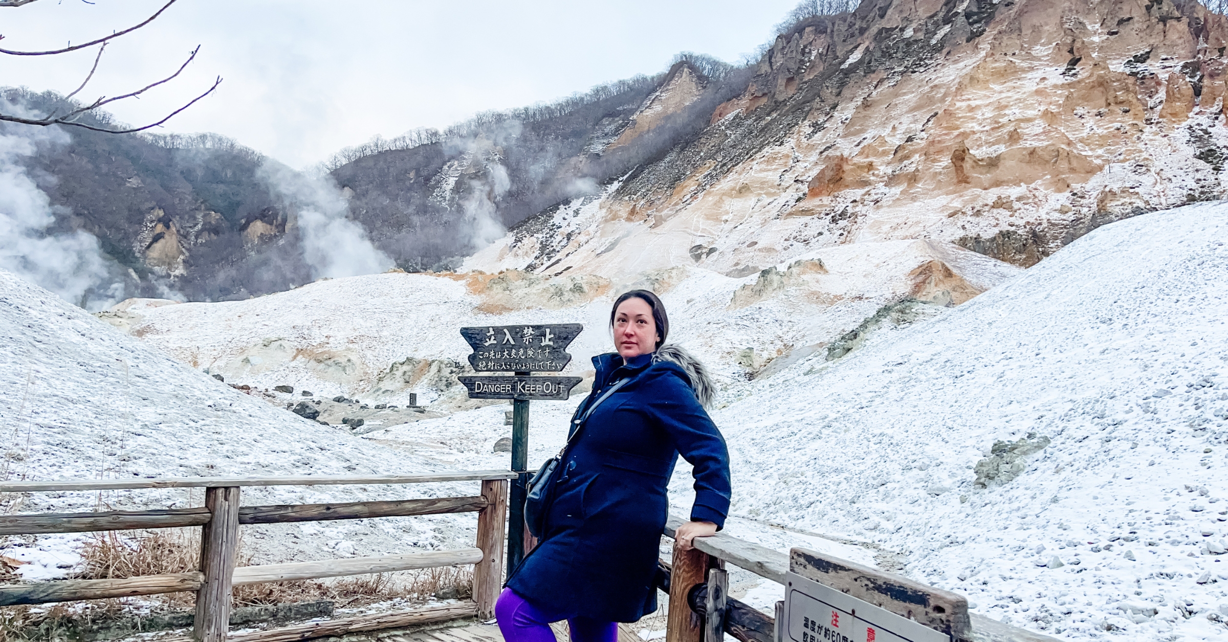A woman standing on a wooden walkway with snow-covered geothermal terrain and steaming hills in the background, next to a sign that reads 'Danger: Keep Out.' Perfect for an article on motivational quotes about struggle and overcoming challenges.