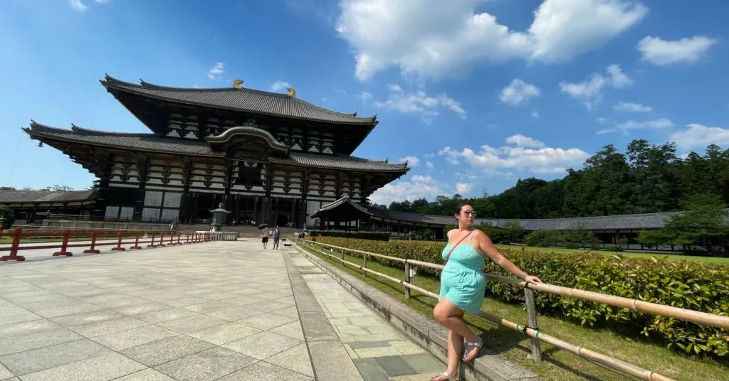 A woman in a bright, serene setting leans against a wooden railing in front of a grand traditional Japanese temple, symbolizing reflection and clarity during a journey of self-discovery. The expansive sky and lush greenery emphasize the vastness of perspective when overcoming an existential crisis. #HowToOvercomeExistentialCrisis