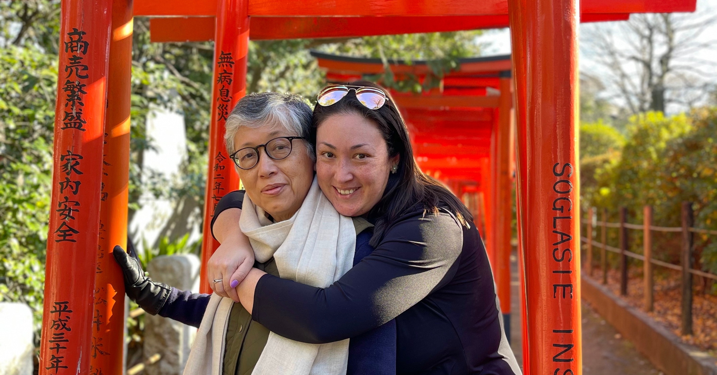 A smiling adult daughter warmly hugs her mother as they stand together amidst vibrant red torii gates in a peaceful outdoor setting. The affectionate gesture and serene backdrop evoke the importance of family connection, making it an ideal representation for discussing how to set healthy boundaries with parents to improve relationships.
