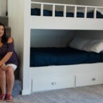 A woman sits by a built-in bunk bed in a neatly arranged, minimalist room, highlighting a cozy and inviting Airbnb setup. This image conveys the appeal of thoughtfully designed spaces in short-term rentals, addressing the question of whether the Airbnb dream is still achievable amidst changing industry trends in 2025.