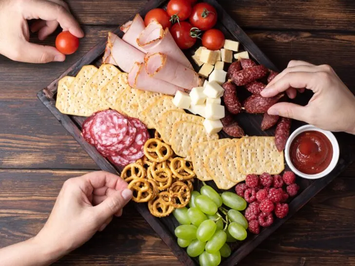 A vibrant charcuterie board featuring a selection of crackers, cured meats, cheese cubes, fruits, and dipping sauce, with hands reaching in to grab snacks. Perfectly illustrates the social allure and millennial obsession with this trendy appetizer style.