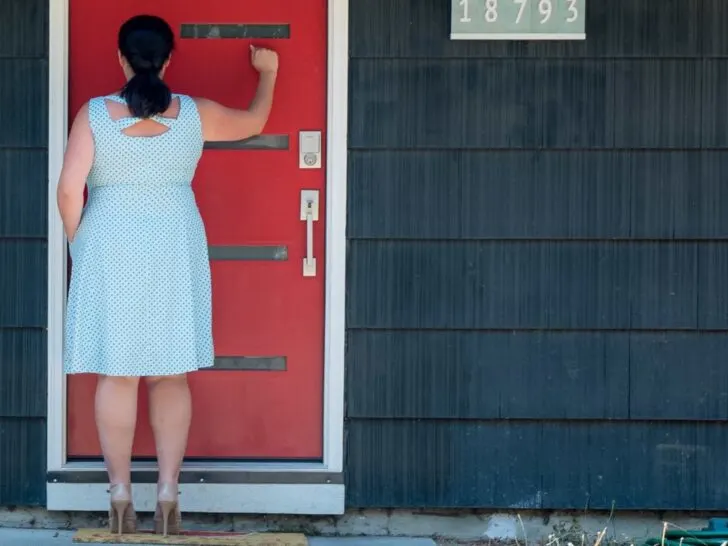 A woman in a polka-dot dress is seen from behind, knocking on a bright red front door with a smart lock installed, emphasizing secure and convenient entry. This image highlights the benefits of using a smart lock for easy and seamless Airbnb guest check-ins, eliminating the need for traditional keys and enhancing the guest experience.