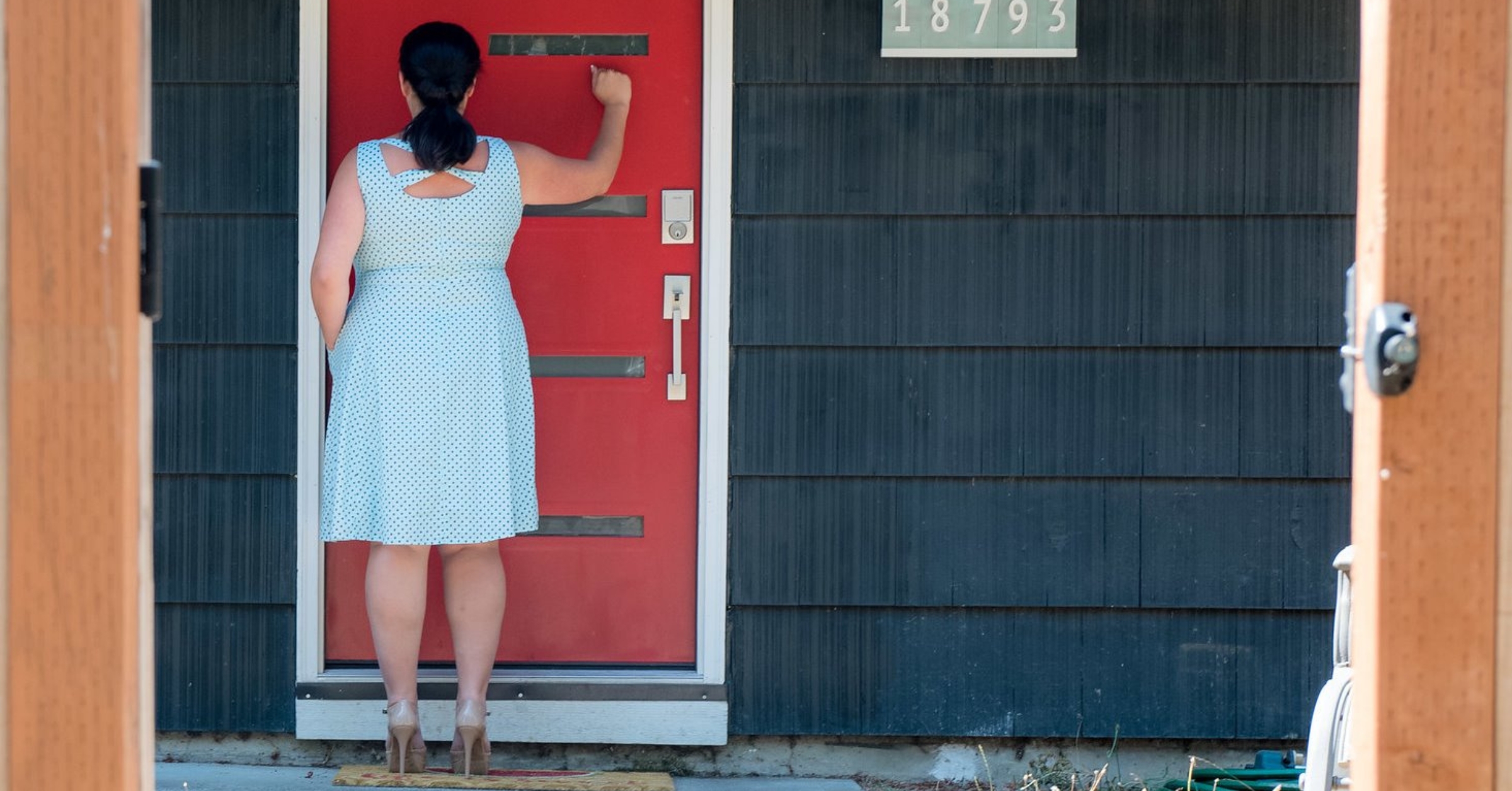 A woman in a polka-dot dress is seen from behind, knocking on a bright red front door with a smart lock installed, emphasizing secure and convenient entry. This image highlights the benefits of using a smart lock for easy and seamless Airbnb guest check-ins, eliminating the need for traditional keys and enhancing the guest experience.
