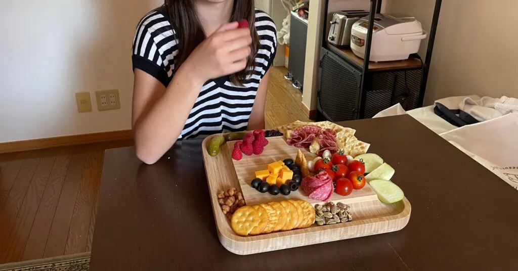 A child enjoys a vibrant charcuterie board featuring crackers, cheddar cubes, black olives, raspberries, salami, cherry tomatoes, pickles, and other finger foods. This image highlights how charcuterie boards have evolved into a family-friendly trend, gaining widespread popularity for their aesthetic appeal and snackable variety. 