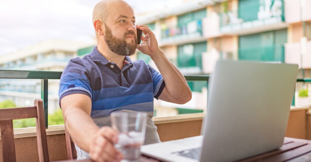 A man sitting on an outdoor balcony, talking on the phone and working on a laptop with a glass of water nearby. This image reflects the flexibility and freedom often associated with outsourcing work and managing virtual assistants remotely.