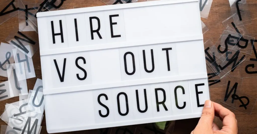 A lightbox sign displaying the words “HIRE VS OUTSOURCE,” with scattered letter tiles around, illustrating the comparison between hiring employees and outsourcing work. This image supports the theme of evaluating different staffing options for business efficiency and cost-effectiveness in an outsourcing guide.
