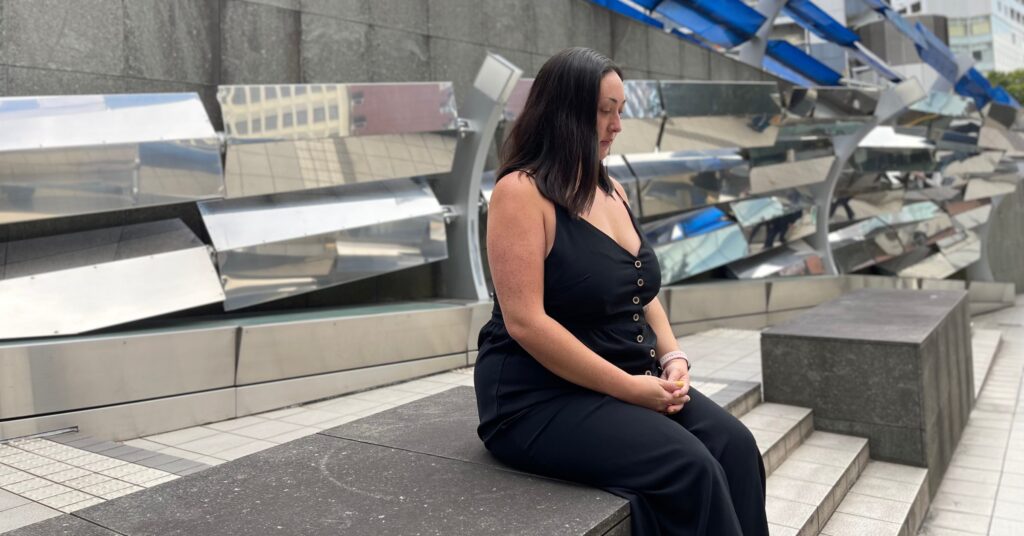 A woman in a black outfit sitting on a concrete ledge in a modern urban environment, with reflective metallic art installations in the background. Ideal for an article on motivational quotes about struggle and perseverance.