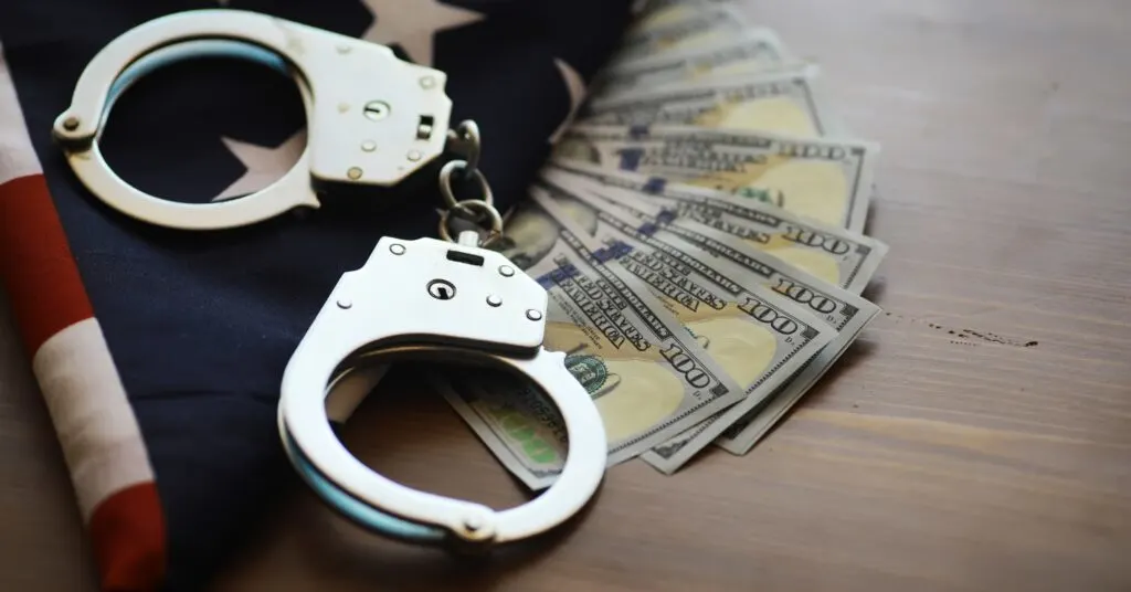 A pair of handcuffs lies next to a stack of U.S. hundred-dollar bills atop an American flag, symbolizing the intersection of law, money, and nationality. This image represents the complexities felons may face when traveling internationally, particularly regarding financial implications and legal restrictions, aligning with the topic of which countries permit entry for travelers with a criminal record.