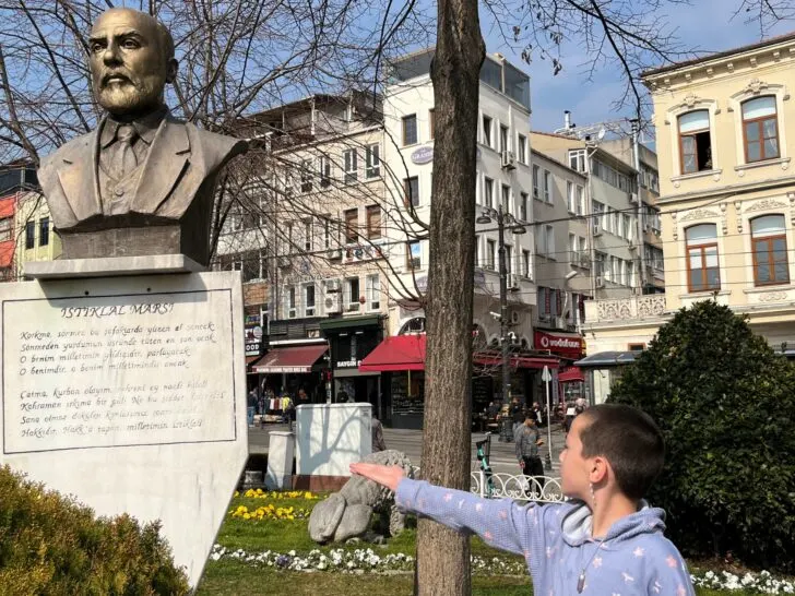 A child pointing at a statue of Mehmet Akif Ersoy in a public square in Istanbul, surrounded by historical buildings and gardens, showcasing educational and cultural things to do in Istanbul, Turkey, with kids.