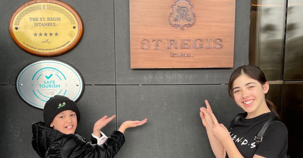 Two children smiling and pointing at the St. Regis Istanbul sign, highlighting one of the best luxury hotels in Istanbul for families, known for its excellent service and family-friendly amenities.