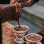 Close-up of traditional Turkish coffee being poured from a hand-engraved copper cezve into two ornate cups with colorful floral designs, illustrating the use of the best Turkish coffee pots for an authentic brewing experience.