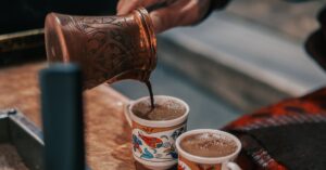 Close-up of traditional Turkish coffee being poured from a hand-engraved copper cezve into two ornate cups with colorful floral designs, illustrating the use of the best Turkish coffee pots for an authentic brewing experience.