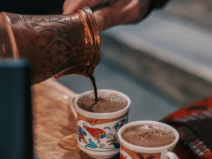 Close-up of traditional Turkish coffee being poured from a hand-engraved copper cezve into two ornate cups with colorful floral designs, illustrating the use of the best Turkish coffee pots for an authentic brewing experience.