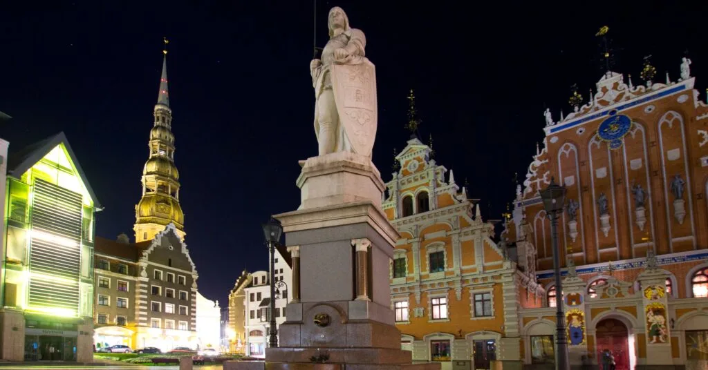 A nighttime view of Riga’s Old Town featuring the Roland statue in the square, with St. Peter’s Church spire and the House of the Blackheads illuminated behind it — an iconic scene reflecting the medieval charm of living in Latvia.