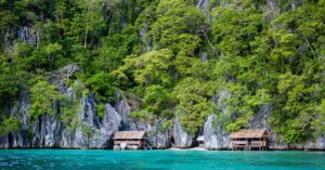 Traditional wooden huts on stilts along the turquoise waters in the Philippines, surrounded by lush green cliffs. A serene tropical escape for those moving to the Philippines.