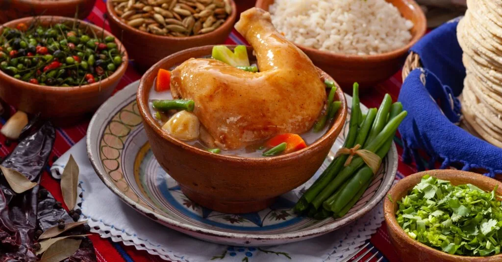 A delicious bowl of traditional Guatemalan caldo de gallina (chicken soup) surrounded by fresh ingredients like green beans, cilantro, and tortillas. A must-try dish when moving to Guatemala and exploring its rich cuisine.