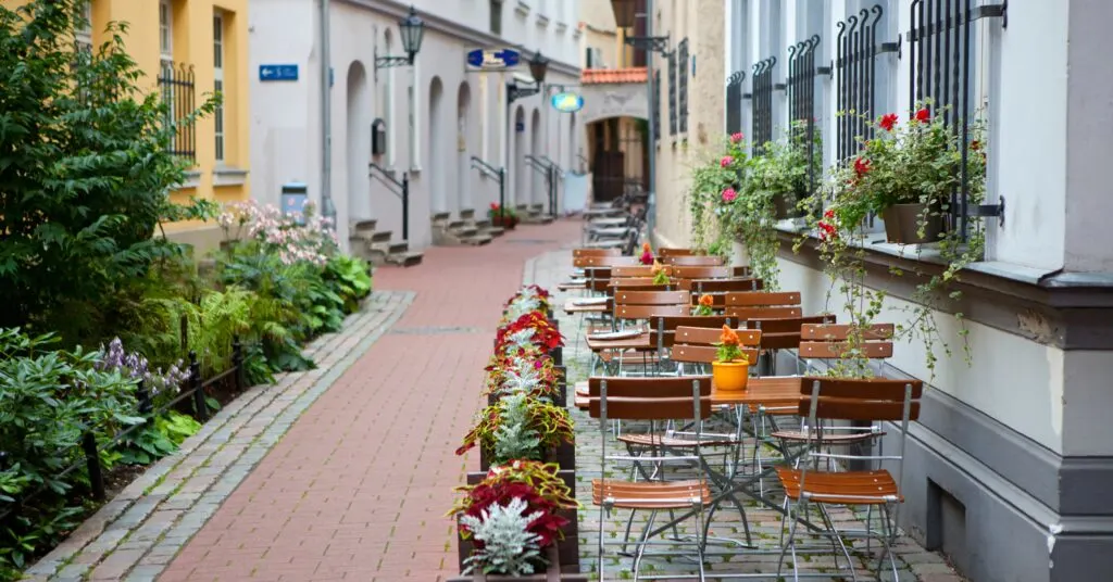 A quiet cobblestone alley in Riga lined with flower boxes, potted plants, and wooden chairs outside a cozy café — showcasing the peaceful, walkable lifestyle of living in Latvia.