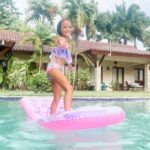 Smiling girl balancing on a pink float in a villa pool surrounded by palm trees, representing playful family moments while living in Dominican Republic.