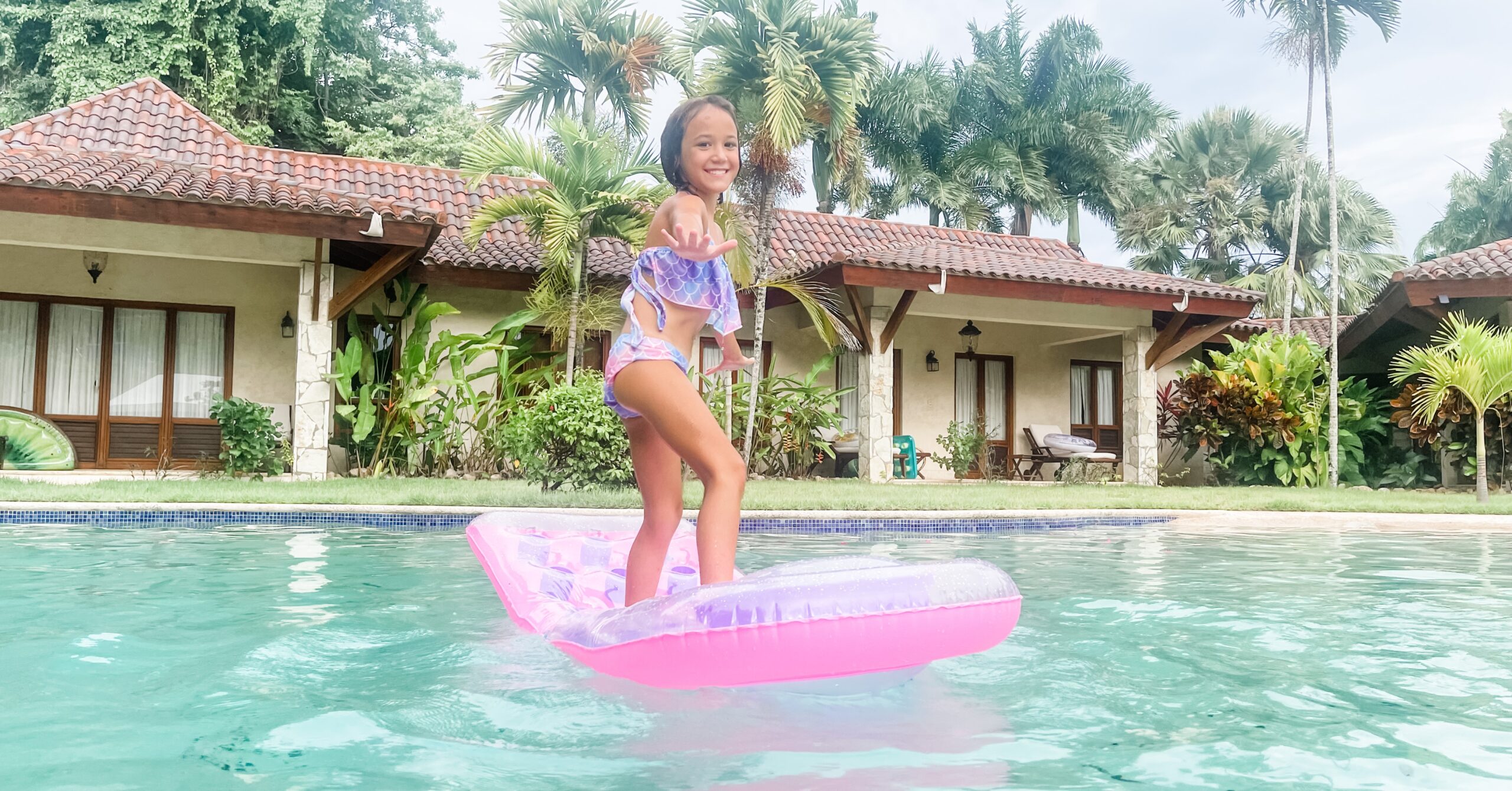 Smiling girl balancing on a pink float in a villa pool surrounded by palm trees, representing playful family moments while living in Dominican Republic.