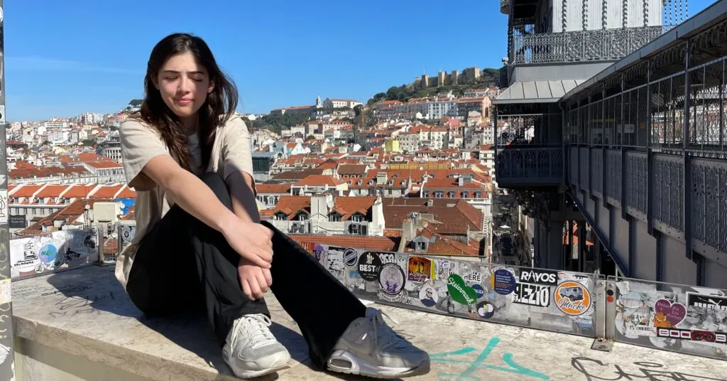 A young woman sits on a ledge enjoying a panoramic view of Lisbon’s terracotta rooftops and historic architecture. Thinking of moving to Portugal? This is the kind of cityscape you could wake up to.