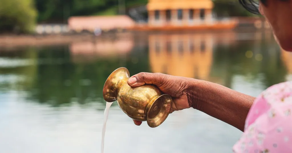 A person pours liquid from a brass vessel into a lake, with a Hindu temple reflecting in the water. Mauritius' rich cultural diversity makes it a unique expat destination.