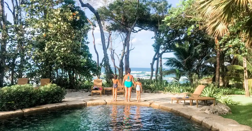 Mother and two daughters holding hands by a lagoon-style pool facing the ocean, illustrating tranquil family time while living in Dominican Republic.