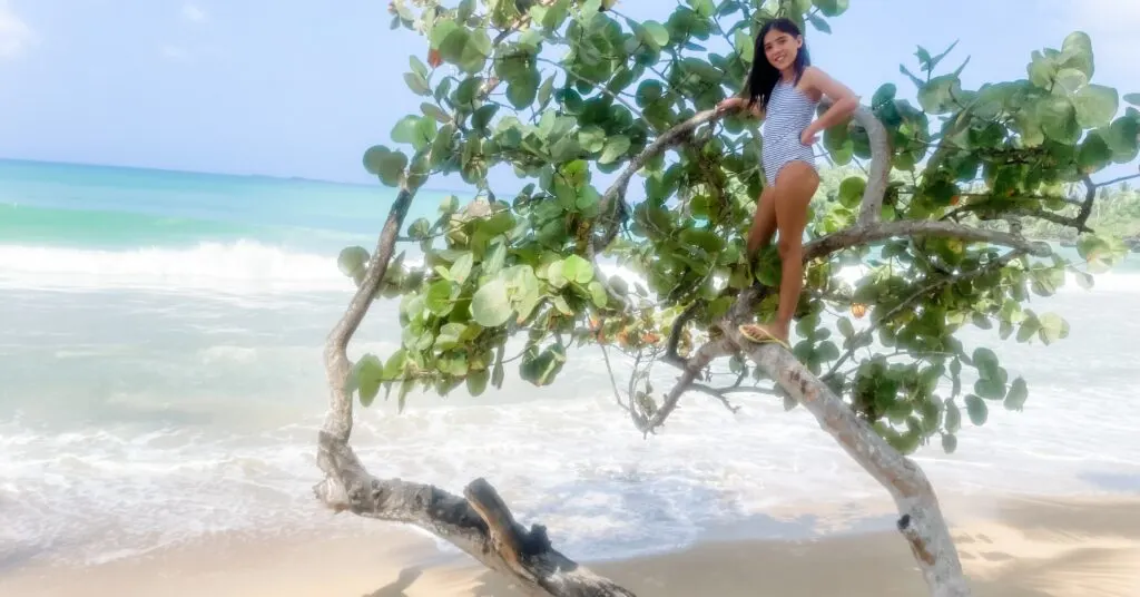 Young girl in a striped swimsuit playfully climbing a seaside tree on a tropical beach, enjoying the natural beauty while living in Dominican Republic.