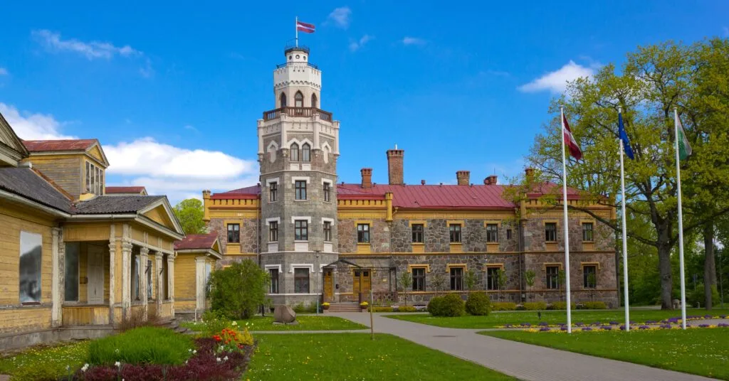 A castle-like building in Latvia flying the national and EU flags, set against a bright blue sky and manicured gardens — a nod to the country’s history and sense of national pride tied to living in Latvia.