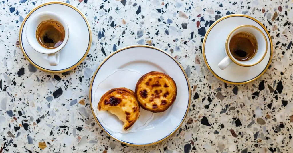 Two golden pastel de nata (Portuguese custard tarts) and two cups of espresso sit on a beautiful speckled table. If you’re thinking of moving to Portugal, these treats might become your daily indulgence.