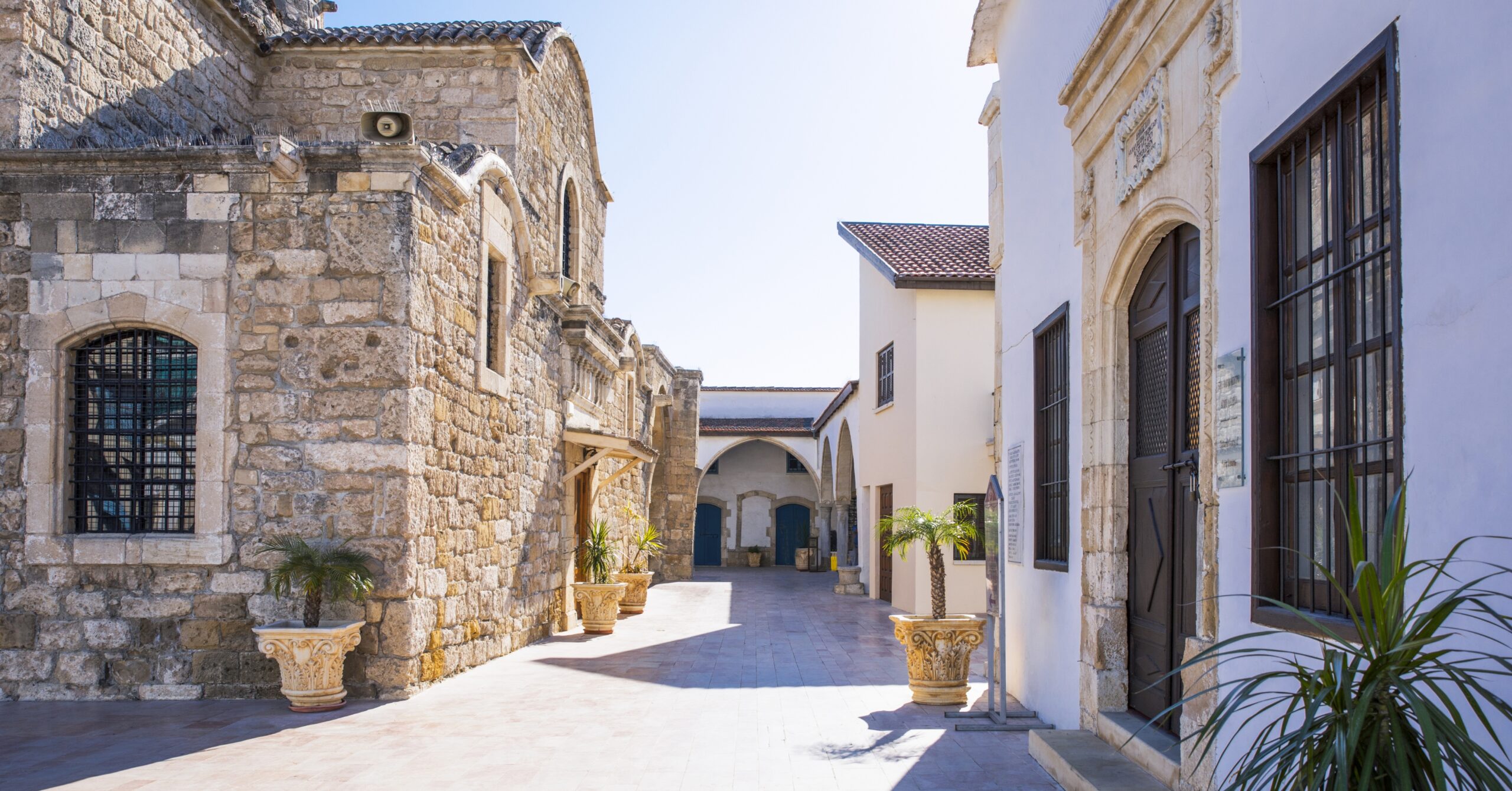A peaceful courtyard surrounded by stone buildings with arched entrances and potted palm trees. The architectural beauty of Cyprus is a major attraction for expats.