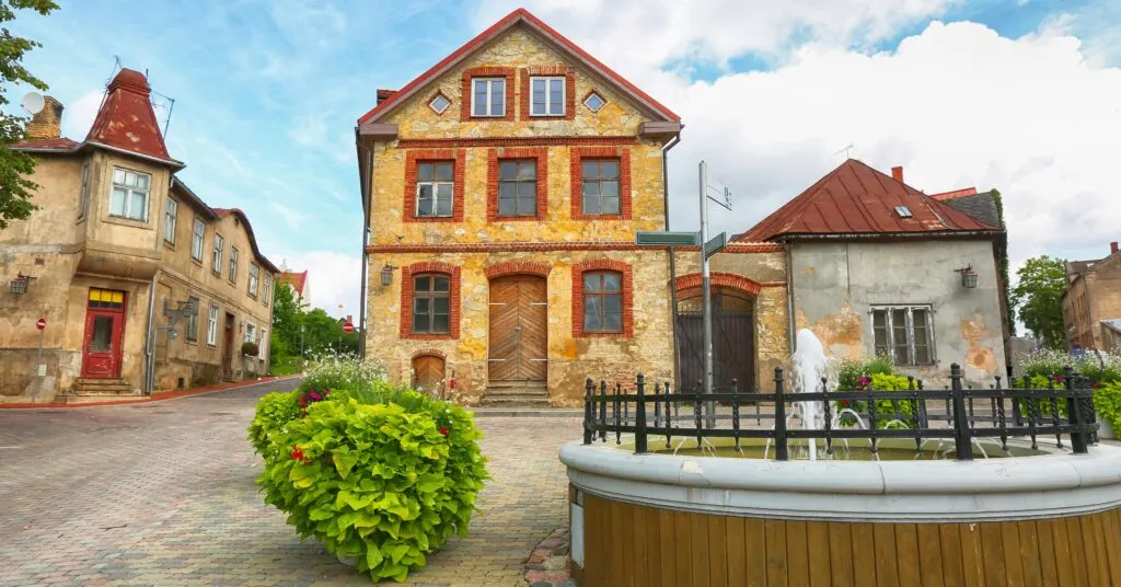 A quaint cobbled street with rustic stone buildings, a wooden fountain in the foreground, and red-roofed houses — illustrating the slower pace of rural living in Latvia.