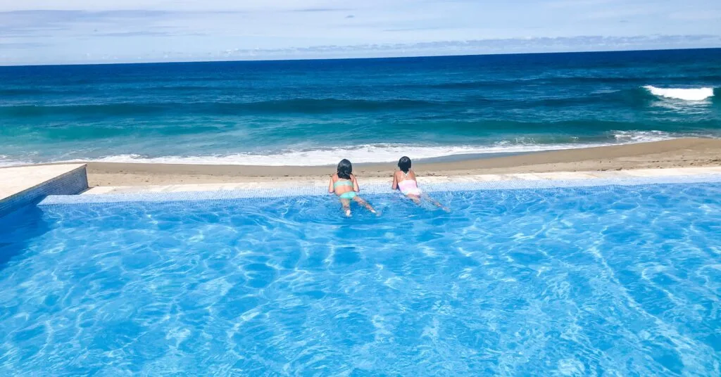 Two young girls lounging at the edge of an infinity pool overlooking a sandy beach and blue ocean—luxury and leisure while living in Dominican Republic.