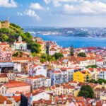 A sweeping view of Lisbon’s red-roofed buildings, with São Jorge Castle on a hill and the Tagus River in the background. If you're thinking of moving to Portugal, Lisbon offers stunning views like this daily.