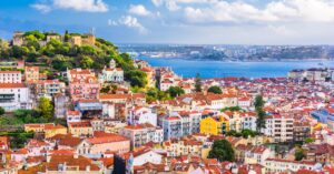 A sweeping view of Lisbon’s red-roofed buildings, with São Jorge Castle on a hill and the Tagus River in the background. If you're thinking of moving to Portugal, Lisbon offers stunning views like this daily.