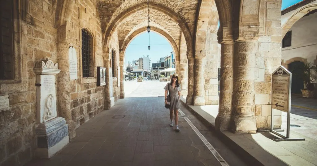 Woman in a summer dress and sunhat walks through an old stone archway toward a sunny plaza, suggesting the charm and walkability of one of the cheapest countries to move for American expats.